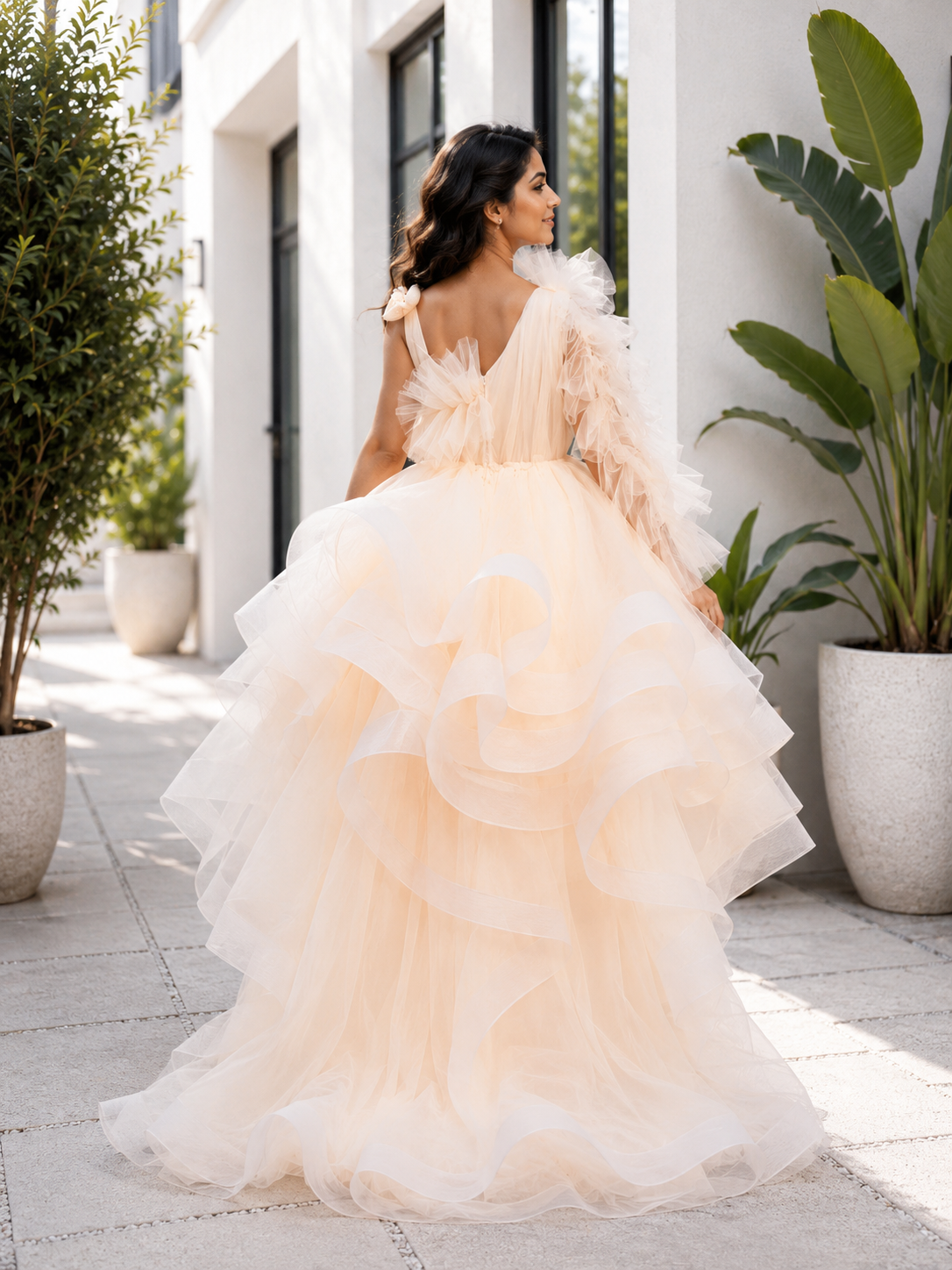 Woman in a peach-colored ruffled gown standing outdoors with plants and a building in the background.
